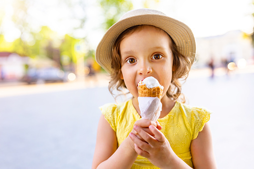 a child eats ice cream in the summer when it's hot outside. Ice cream in a waffle cone. A happy and contented child in a hat and dress.