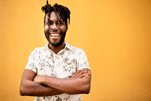 Portrait of a young man laughing while standing with his arms crossed outside in front of a yellow wall