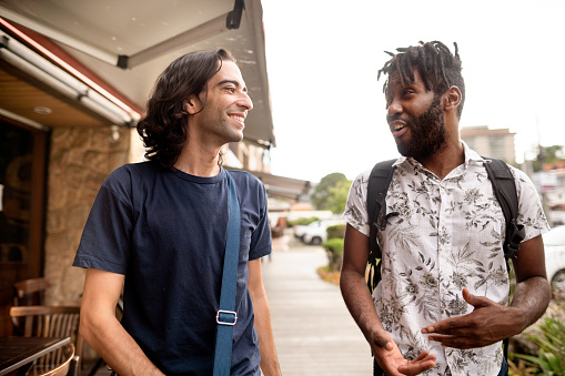 Two diverse young male friends talking together and smiling while walking along a sidewalk in the city