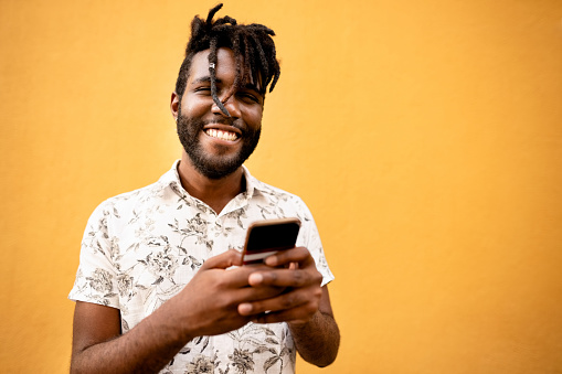 Portrait of a stylish young african man with his cell phone standing against orange background looking at camera and smiling