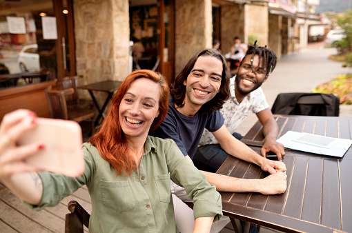 Diverse group of smiling friends talking a selfie while sitting together outside at a sidewalk cafe table