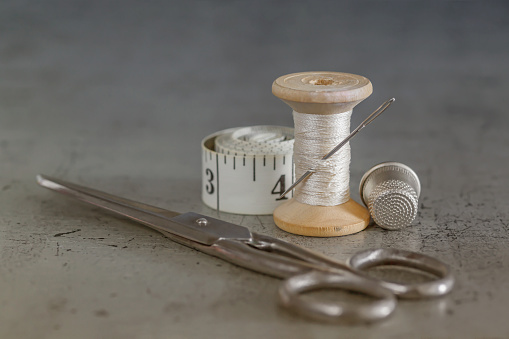 Hand sewing tools, threads on a wooden spool, sewing needle with scissors, thimble and tailor's tape on an old surface. Sewing thread background. Retro style. Selective focus