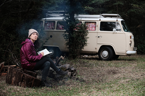 Charming woman in jacket and cap writing in her diary sitting near a campfire and her van in the middle of the forest.