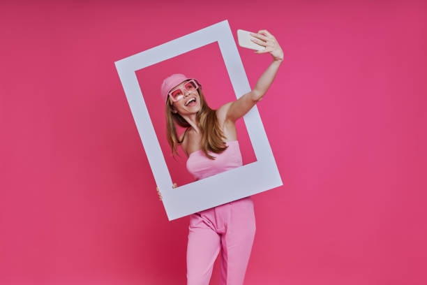 Playful young woman making selfie and looking through a picture frame while standing against pink background stock photo