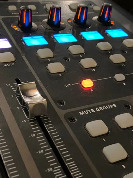 black soundboard with teal lights. Slider knobs to increase and decrease sound levels. Gray buttons, red and black knobs, and a small round orange light.