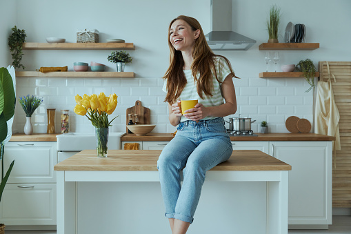 Happy young woman holding yellow cup and smiling while sitting on the kitchen counter