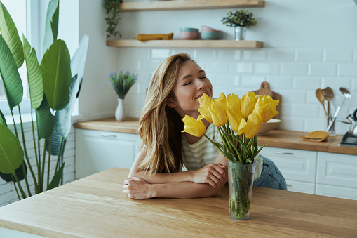 Happy young woman leaning at the kitchen desk and smelling yellow tulips