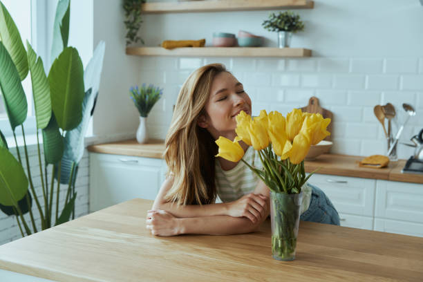 Happy young woman leaning at the kitchen desk and smelling yellow tulips stock photo