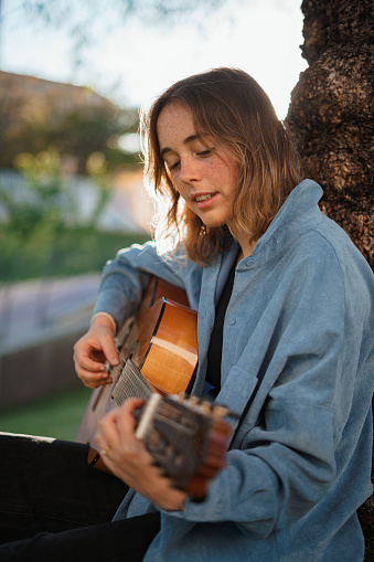 Smiling girl playing guitar leaning against tree in park.