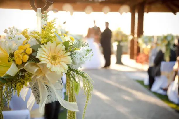 outdoor wedding, center aisle with flower bouquet hanging on hook. Flowers are yellow and white. Bride and groom are out of focus in the background. Focus is on flower bouquet