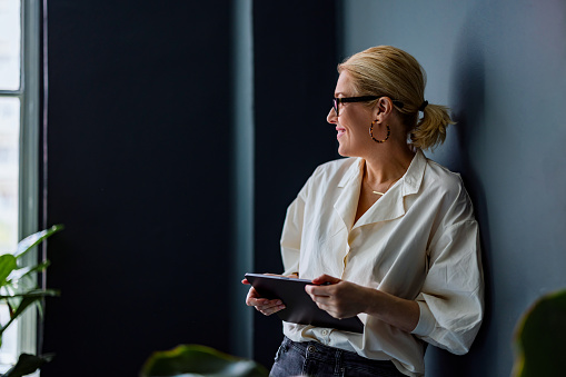 Smiling businesswoman holding digital tablet while looking trough window and standing in her office.