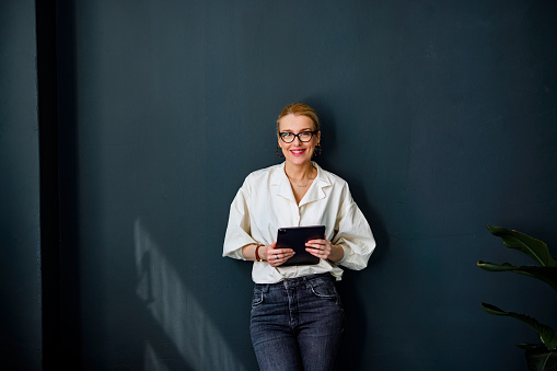 Happy businesswoman holding a tablet while standing in front of the black wall and looking at camera.