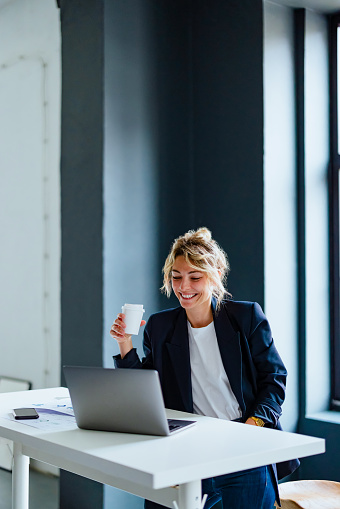 Happy businesswoman holding cup of coffee while reading business report on her laptop computer.