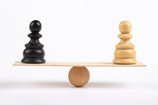 White and a black pawn chess piece stand on wooden seesaw on white background