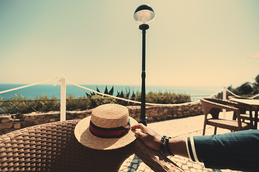 View of the hand of an elegant black man wearing a fashionable tailored navy blue suit and holding a classic golden straw hat while sitting in an outdoor coastal restaurant on a warm sunny day