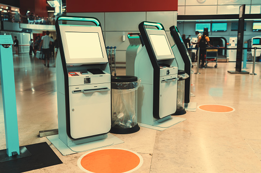 Side view with mock-ups of blank white LCD screens of futuristic electronic ticket terminals or self-service check-in machines in a modern airport or a train station depot; social distancing stickers