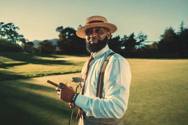 An African golfer on a golf field A portrait of a dashing mature bearded black man in a fashionable outfit with a straw hat and braces standing on a golf field in the evening with a sunset, holding a golf club and a cigar in the hands vintage cigar stock pictures, royalty-free photos & images
