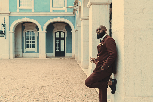 A dapper handsome African American bald bearded guy in a dark-red tailored costume is leaning against the wall, looking away and holding eyeglasses in his hand; a copy space place on the left
