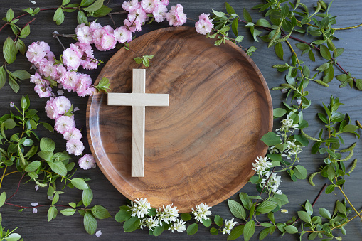 Frame of branches with pink and white spring flower blossoms and green leaves around a Christian cross on a circular teak wood tray all on a dark wood background with copy space shot from above