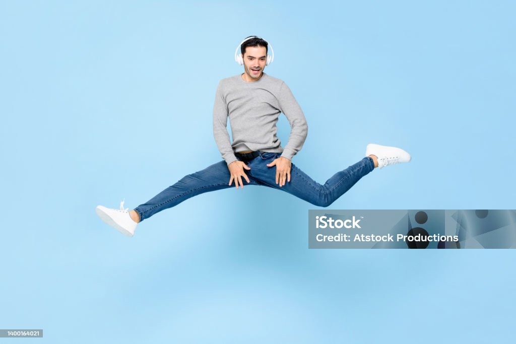 Fun energetic young Caucasian man wearing headphones listening to music and jumping with spread legs in studio light blue color isolated background Doing the Splits Stock Photo