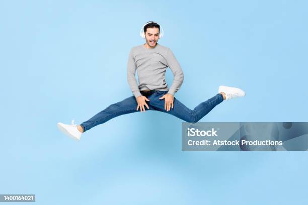 Fun Energetic Young Caucasian Man Wearing Headphones Listening To Music And Jumping With Spread Legs In Studio Light Blue Color Isolated Background Stock Photo - Download Image Now