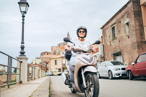 Mujer alegremente sonriente con casco y gafas de sol montando rápidamente el moto scooter en las calles del casco antiguo siciliano en forza d'Agro. Feliz concepto de vacaciones y transporte italiano. photo