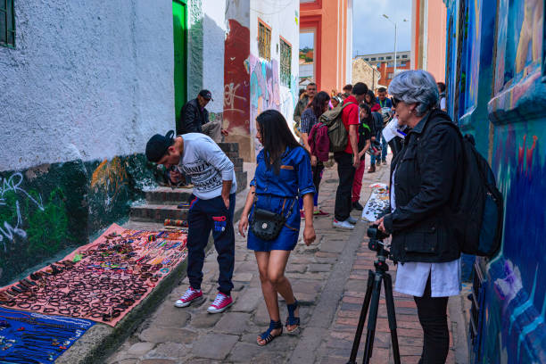 Bogota, Colombia - Tourists And Local Colombians On The Calle del Embudo, In The Historic La Candelaria District Of The Andes Capital City In South America. Bogota, Colombia - July 20, 2016: Local Colombian checking out trinkets and handicraft on the narrow Calle del Embudo one of the most colorful streets in the historic La Candelaria district of Bogotá, the Andean capital city of the South American country of Colombia. The street leads to the Chorro de Quevedo, the plaza where it is believed the Spanish Conquistador, Gonzalo Jiménez de Quesada founded the city in 1538. Many street facing walls in this area are painted with either street art or the legends of the pre-Colombian era, in the vibrant colours of Colombia. The altitude at street level is 8,660 feet above mean sea level. Photo shot on an overcast morning; horizontal format. calle del embudo stock pictures, royalty-free photos & images