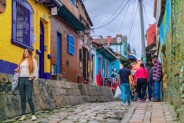 Bogota, Colombia - Tourists And Local Colombians On The Calle del Embudo, In The Historic La Candelaria District Of The Andes Capital City In South America. Bogota, Colombia - October 20, 2019: Both Tourists and local Colombian people walk up the narrow Calle del Embudo one of the most colorful streets in the historic La Candelaria district of Bogotá, the Andean capital city of the South American country of Colombia. The street leads to the Chorro de Quevedo, the plaza where it is believed the Spanish Conquistador, Gonzalo Jiménez de Quesada founded the city in 1538. Many street facing walls in this area are painted with either street art or the legends of the pre-Colombian era, in the vibrant colours of Colombia. The altitude at street level is 8,660 feet above mean sea level. Photo shot on an overcast morning; horizontal format. calle del embudo stock pictures, royalty-free photos & images