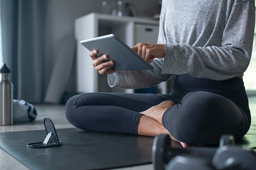 Sporty young Asian woman exercising at home, watching fitness video on Internet or having online fitness class, using tablet , living room interior, copy space