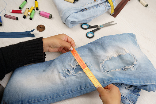 measuring jean pants with tape measure, work table with sewing instruments such as colored threads, scissors, craft occupation and walppaper, studio and objects
