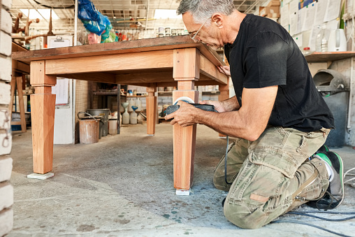 Senior male carpenter sanding wooden table with electric sander in workshop