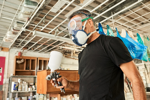 Senior male carpenter wearing protective googles and respirator with wood staining spray gun in his workshop