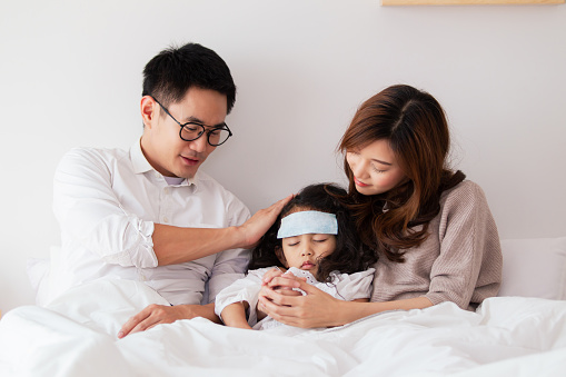 Asian sick girl sleeping on the bed with fever gel pack on her forehead. Father and mother take care of their little young daughter who got a fever from flu virus. sick little kid laying on the bed.