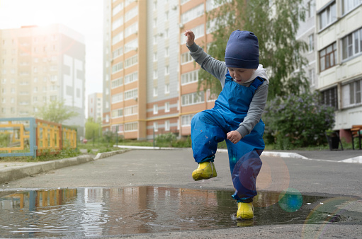 A little boy in rubber boots stomps on puddles in the courtyard of a multi-storey building. Summer in the city.