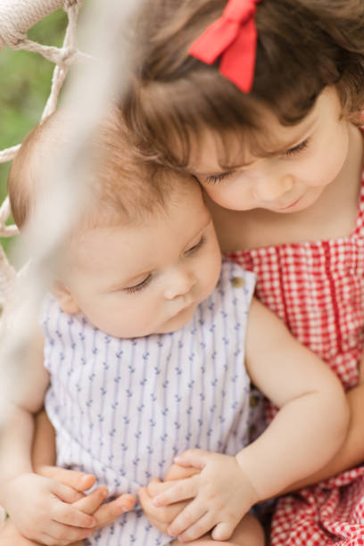 A 5-Month-Old Baby Boy Being Held & Snuggled By His 3-Year-Old Big Sister While They Swing on a Boho Outdoor Chair Swing Hanging From a Large Tree in South Florida for a Festive & Patriotic Memorial Day Celebratory Picnic on a Hot Summer Day. Children Wearing Red, White & Blue clothing. un bebé de 5 meses de edad sostenido y acurrucado por su hermana mayor de 3 años mientras se balancean en un columpio de silla boho al aire libre colgando de un gran árbol en el sur de la florida para un picnic festivo y patriótico de celebración del