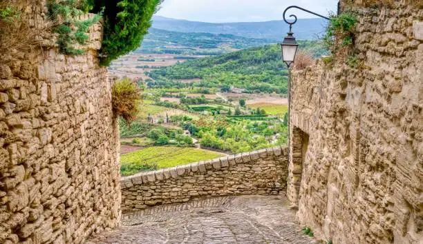 Focus on stone walls and a cobblestone street of the medieval village of Gordes in the Luberon Region of Provence, France, overlooking a wide valley of cultivated fields, orchards and vineyards below.