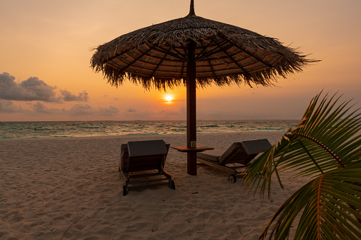 Beautiful scenic view of the sunset from the shore with two sun loungers and a straw umbrella in the foreground. Beach resort relaxation vacation concept.