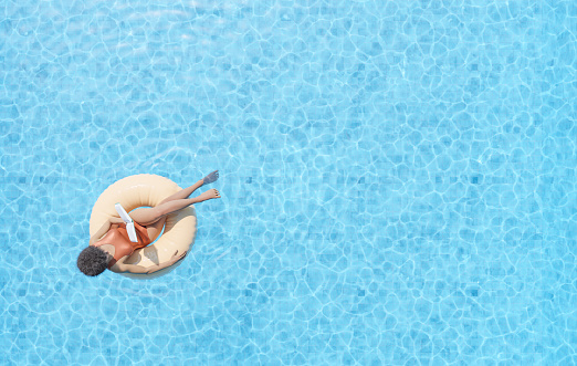 Anonymous ethnic lady floating on rubber ring in swimming pool and reading book