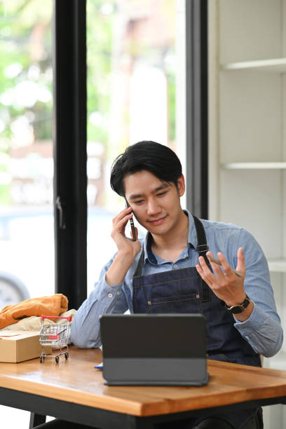 istock Young small business owner using computer and talking on mobile phone for confirming orders from customer. Online selling, e-commerce concept. 1400203634