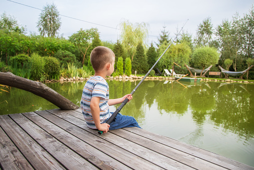 A small European boy in a striped T-shirt, sitting with his back. The child is fishing with a long rod. Sunny summer day. The concept of childhood, happiness and tranquility. June 1st,