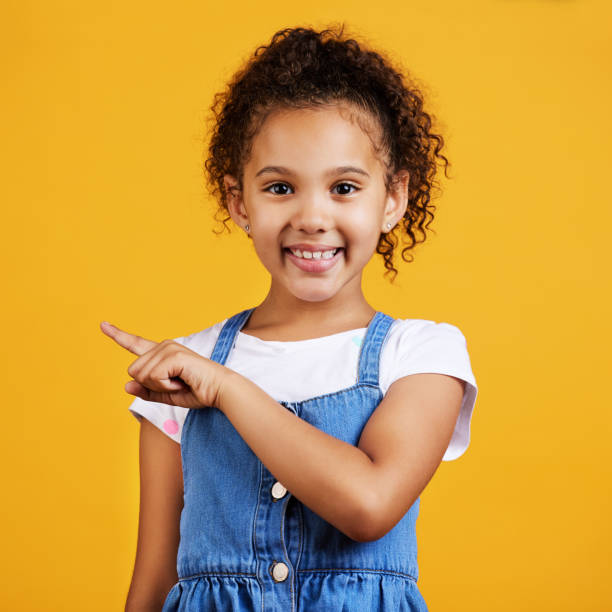 istock Studio portrait mixed race girl pointing sideways towards copyspace isolated against a yellow background. Cute hispanic child posing inside. Happy and cute kid showing or endorsing company or product 1400209703