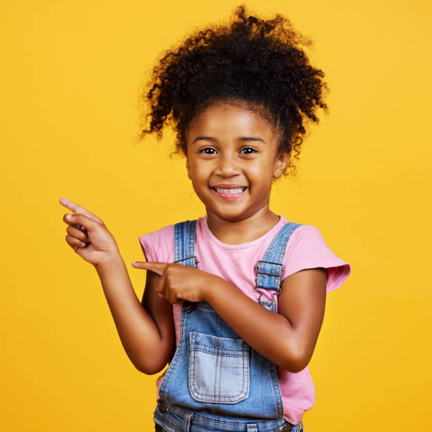 istock Studio portrait mixed race girl pointing sideways towards copyspace isolated against a yellow background. Cute hispanic child posing inside. Happy and cute kid showing or endorsing company or product 1400209870