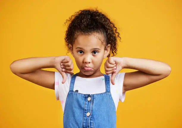 Studio portrait mixed race girl giving thumbs 
down isolated against a yellow background. Cute hispanic child posing inside. Unhappy and upset kid being negative and saying, I disagree or disapprove