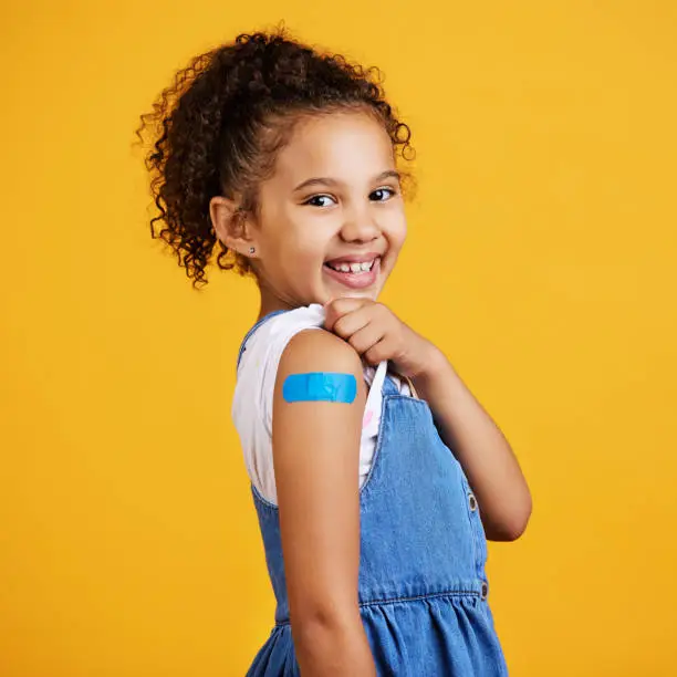 Studio portrait mixed race girl showing a plaster on her arm Isolated against a yellow background. Cute hispanic child lifting her sleeve to show injection site for covid or corona jab and vaccination