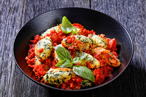 Malfatti, Italian spinach ricotta dumplings  in tomato sauce with herbs and grated parmesan cheese in black bowl on dark wooden table