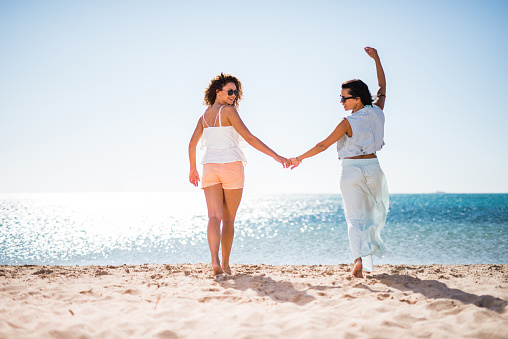 Rear view of happy women having fun while holding hands and running in summer day on the beach. Copy space.