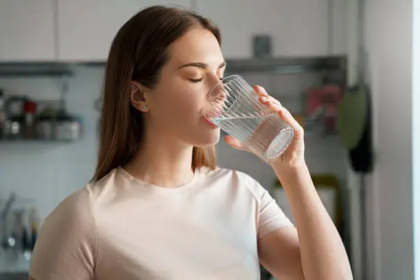Thirsty young woman drinking fresh water from glass. Home office kitchen interior. Headshot portrait. Dehydration prevention, normal bowel function and balance of body maintenance concept