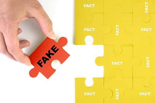 The hand holds a red puzzle with the inscription fake next to the assembled composition of yellow puzzle facts. The concept of substitution of facts in the media.