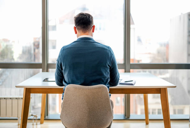 Rear view of businessman working at desk in front of windows at office Rear view of businessman working at desk while sitting in front of windows at corporate office man in the desk back view stock pictures, royalty-free photos & images