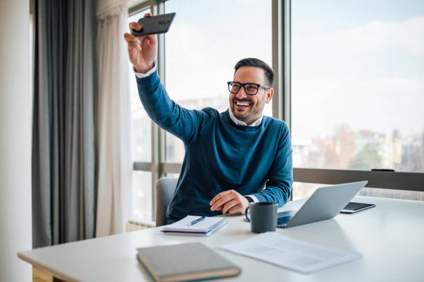 Happy businessman taking selfie on smart phone while working at office desk